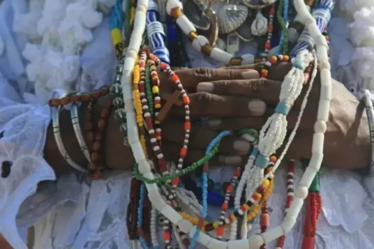 Imagem mostra a mão de uma mullher negra em volta de fio de contas, brajá, · guias e colar de santo típicos do candomblé.
