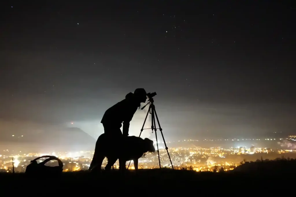 Homem obseva o espaço com um telescópio em uma serra acompanhado de um cachorro